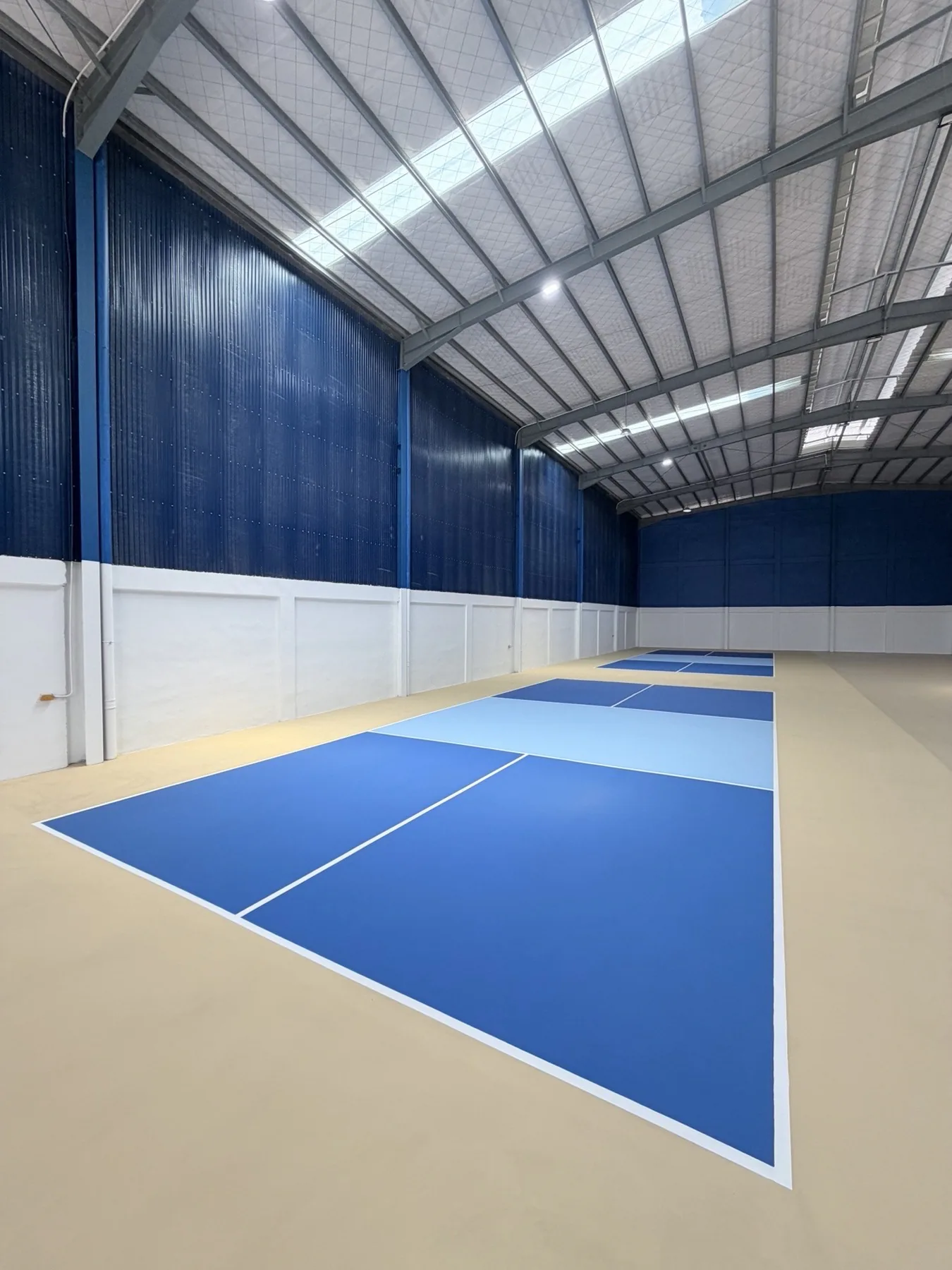 An indoor blue pickleball court beneath a high steel roof